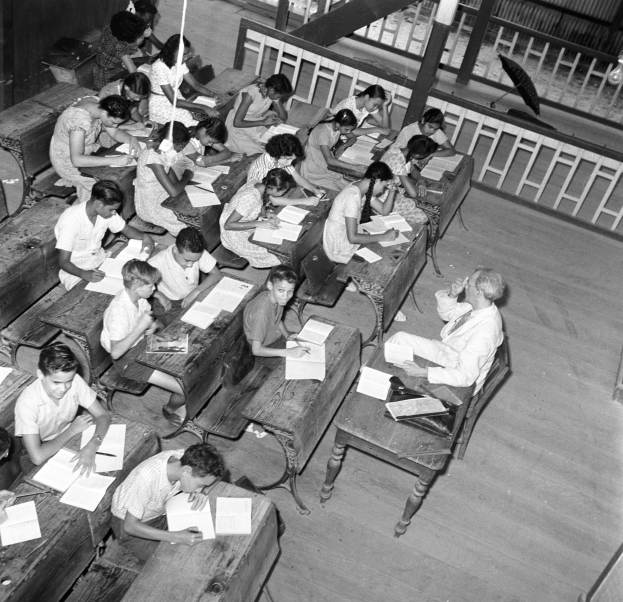 Ein Schwarz-Weiß-Foto einer Klasse mit Kindern an Tischen, Büchern und Gegenständen auf den Tischen, einem Geländer und einem Schirm im Hintergrund.