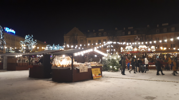 Ein nächtlicher Weihnachtsmarkt in einer verschneiten Straße mit Menschen, Kinderwagen, beleuchteten Hütten, Pflanzen, Bäumen, Gebäuden und Schildern unter einem bewölkten Himmel.