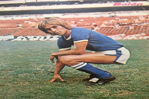 Ein Mann kniet auf einem Fußballfeld, umgeben von sitzenden Menschen mit Stühlen und Tafeln mit Text, die Schuhe tragend.