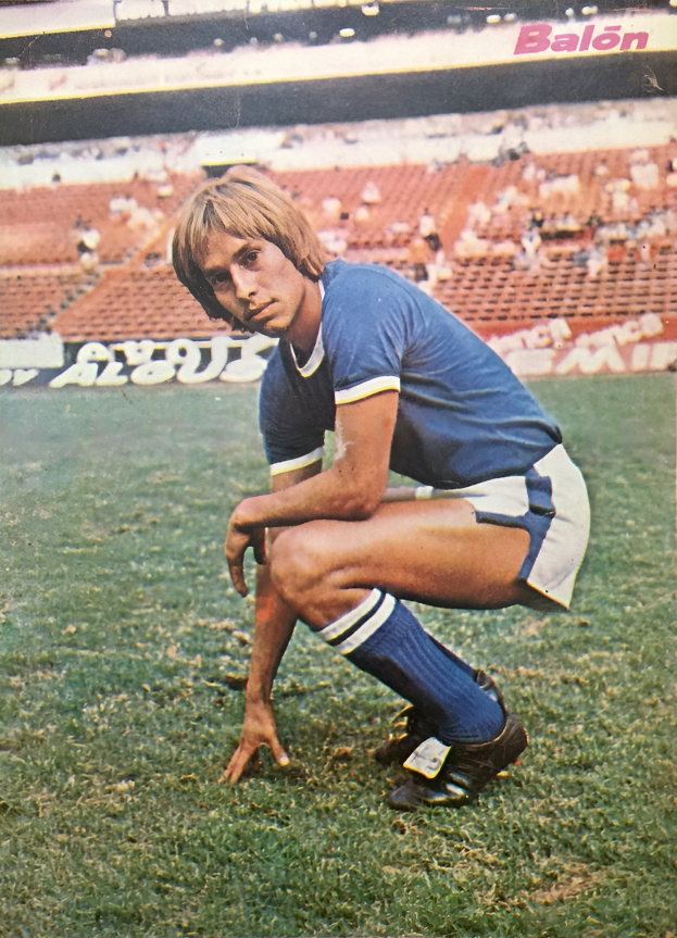 Ein Mann kniet auf einem Fußballfeld, umgeben von sitzenden Menschen mit Stühlen und Tafeln mit Text, die Schuhe tragend.