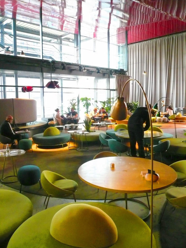 Ein geräumiger Hotel-Salon mit grünen Stühlen, Tischen, Sofas, stehenden Menschen, Pflanzen, einer Stehlampe, Vorhängen und Deckenleuchten.