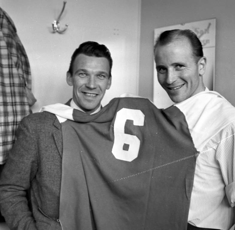 Zwei Männer in Schwarz-Weiß mit strahlenden Gesichtern, die ein Fußballtrikot hochhalten, mit einer Tafel und einem Bügel an der Wand dahinter.