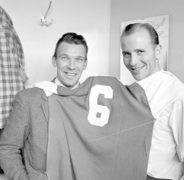 Zwei Männer in Schwarz-Weiß mit strahlenden Gesichtern, die ein Fußballtrikot hochhalten, mit einer Tafel und einem Bügel an der Wand dahinter.