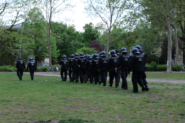 Eine Gruppe von Polizisten in schwarzen Uniformen und Helmen geht über ein grünes Feld, mit Fahrrädern, Laternenmasten, Pflanzen, Bäumen, Gebäuden und einem klaren blauen Himmel im Hintergrund.
