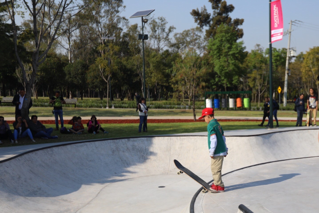 Ein junger Junge in einem grünen T-Shirt, braunen Hosen und einer roten Kappe fährt auf einem Skateboard eine Rampe auf einem Skatepark hinunter, mit Menschen, Bäumen und Laternen im Hintergrund.