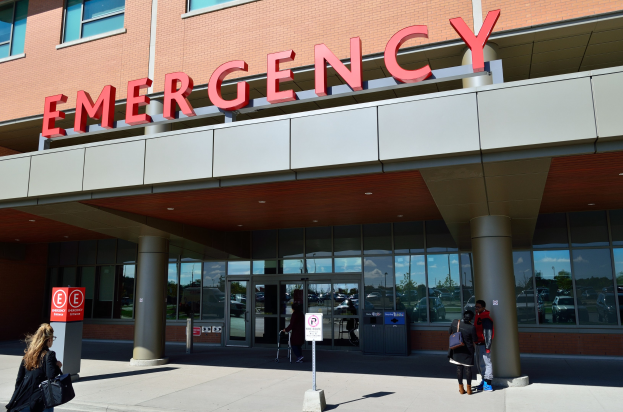 Eine Gruppe von Menschen vor einem Notfallgebäude mit Glaswänden, Säulen, einem Namensschild, Hinweistafeln auf Pfählen und einem Mülleimer.