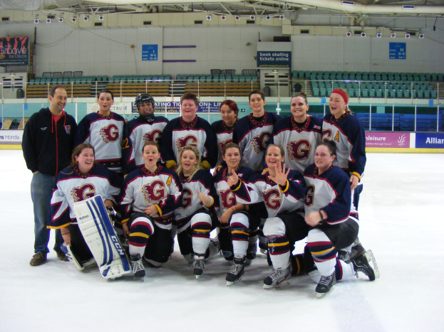 Eine Gruppe von Frauen-Eishockeyspielerinnen in Helmen und Schlittschuhen posiert für ein Foto auf dem Eis, eine hält einen Hockey-Schläger, mit Stühlen, Tafeln mit Text, Geländern und Deckenleuchten im Hintergrund.