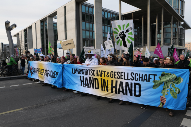 Eine Gruppe von Menschen, die eine Fahne mit der Aufschrift "Landwirtschaft und Gesellschaft Hand in Hand" tragen, geht eine Straße entlang, mit Gebäuden, Bäumen und Polen im Hintergrund unter einem klaren blauen Himmel.