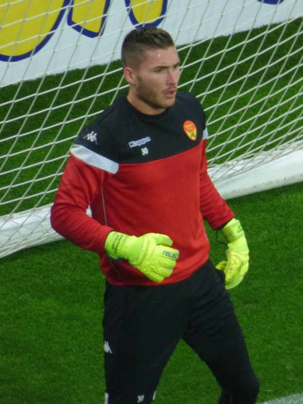 Ein Mann in rotem T-Shirt und schwarzen Hosen steht auf einem Fussballfeld mit einem Netz und einer Fahne im Hintergrund, trägt Fussballhandschuhe.
