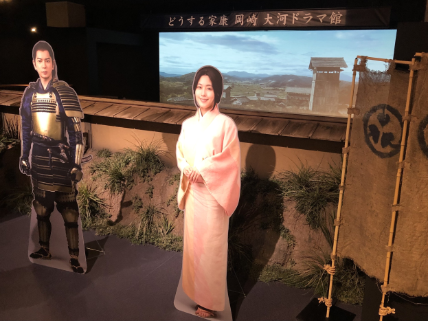 Zwei Menschen in traditioneller japanischer Kleidung - eine Frau in einem rosafarbenen Kimono und ein Mann in Samurai-Rüstung - lächeln gemeinsam in einer Szene aus einem japanischen Film, mit Pflanzen, einem Bildschirm und Beleuchtung im Hintergrund.
