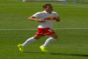 Ein professioneller Fußballspieler in einem weißen Hemd und roten Shorts, der auf einem Feld mit einem Tor im Hintergrund und einigen Menschen in der Nähe läuft.