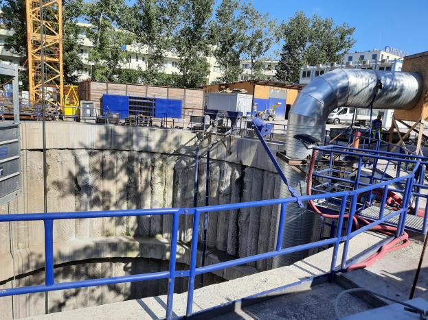 Eine Baustelle auf einer Kläranlage mit einem großen Metallrohr in der Mitte, umgeben von Geländern, Rohren, Maschinen und anderen Gegenständen, mit Gebäuden, Bäumen und einem Turm im Hintergrund.