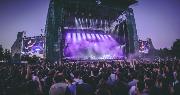 Eine große Menschenmenge versammelt sich auf einem Musikfestival mit einer Bühne, auf der Musiker spielen, die von Lichtern beleuchtet und von Bildschirmen umgeben sind, vor einer Kulisse aus einem Gebäude, Bäumen und einem klaren blauen Himmel.