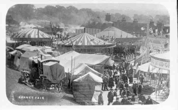 Ein altes Schwarz-Weiß-Foto einer lebhaften Karnevals-Szene mit Zelten, Menschen, Karren, Pfählen, Bäumen und Gebäuden im Hintergrund, beschriftet mit "Barnet Fair, London, England".