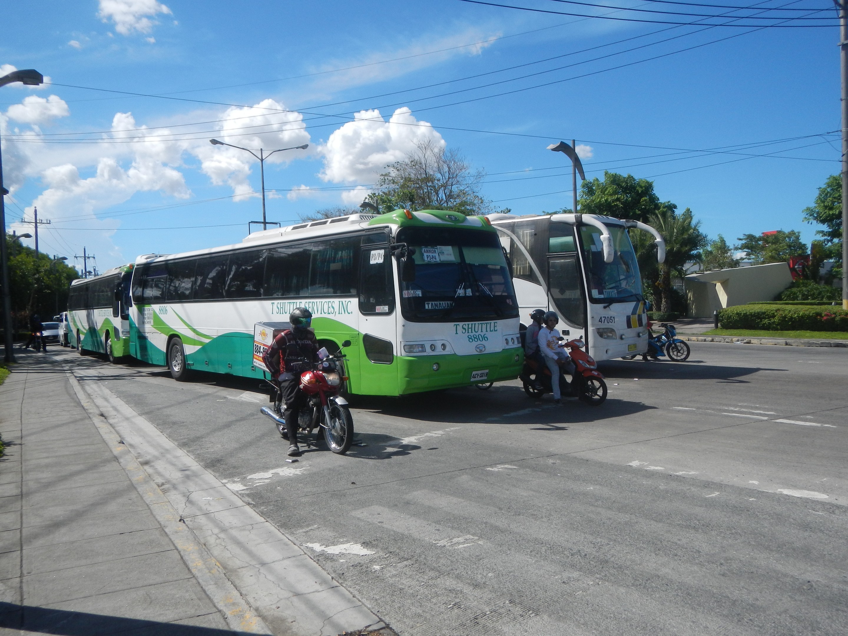 Grüner und weißer Shuttlebus am Straßenrand geparkt mit Motorrädern davor, Fußweg mit Gras und Pflanzen links und im Hintergrund Gebäude, Bäume, Laternenpfähle und einen klaren blauen Himmel.
