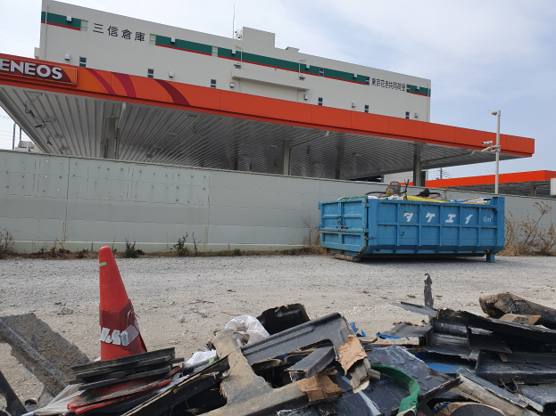 Ein großes Gebäude mit erheblichen Rückständen, darunter Schrottmetall, einen Verkehrskegel und einen blauen Container davor, mit Pflanzen, Polen und dem Himmel im Hintergrund, was auf Abrissarbeiten hinweist.