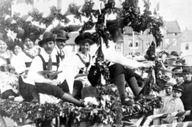 Eine Gruppe von Menschen in traditioneller bayrischer Tracht fährt auf einem mit Laub geschmückten Wagen in einem Umzug mit, einige spielen Musikinstrumente, mit Gebäuden und einem klaren Himmel im Hintergrund, dargestellt in Schwarz-Weiß.