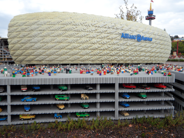 Ein großes Lego-Modell des Allianzstadions in München, Deutschland, mit Menschen davor, umgeben von Bäumen, Pfählen, einem Turm und Pflanzen, unter einem bewölkten Himmel.