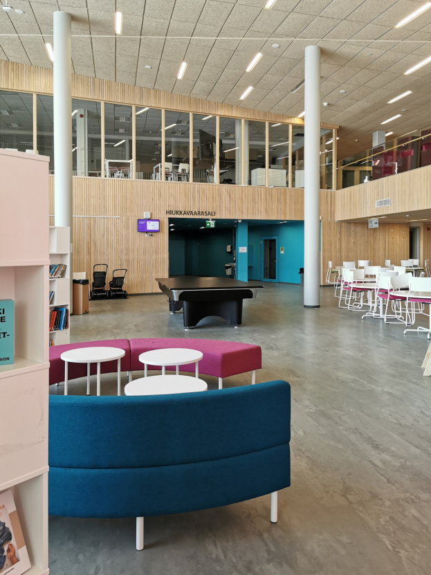 Ein geräumiger Raum, der einer Schulbibliothek ähnelt, mit Tischen, Stühlen, Sofas, Hockern, Bücherregalen, Säulen, Deckenleuchten und einem Wandfernseher.