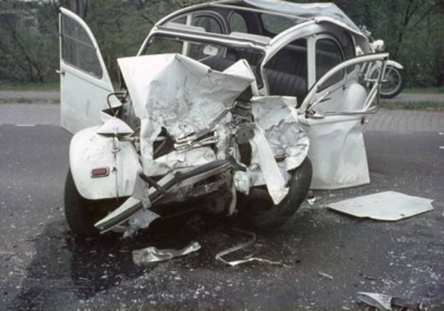 Schwerbeschädigte Kfz am Stra√enrand mit Frontalaufprall, Bäume und ein Gebäude im Hintergrund, weiteres Fahrzeug in der Ferne sichtbar.