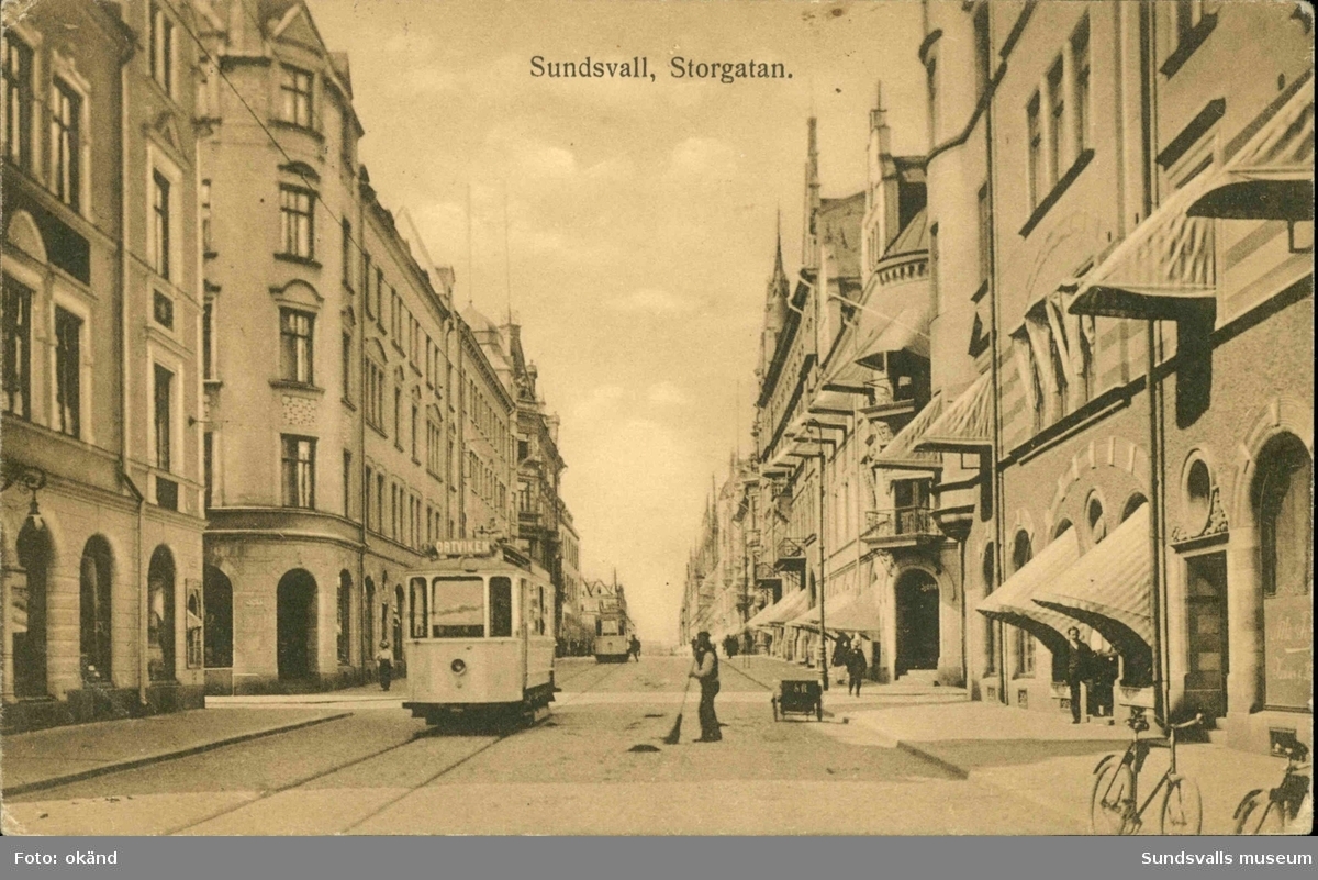 Ein altes Foto einer Stadtstraße mit einer Tram, Gebäuden mit Fenstern, Menschen, die gehen, Fahrrädern und einem bewölkten Himmel, mit Text oben und unten auf dem Bild.