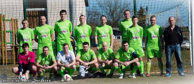 Eine Gruppe von Männern in grünen T-Shirts steht und hockt auf einem Fußballfeld mit einem Ball vor ihnen, ein Torpfosten rechts und ein Hintergrund aus Bäumen, Gebäuden, Pfählen, Fahrzeugen und dem Himmel; Text unten links liest "Bundesliga 1-2-3-4-5-6-7-8-9-10-11-12-13-14-15-16-17-18-19-20-21-22-23-2