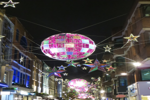 Eine festlich geschmückte Stadtstraße bei Nacht mit vielen Lichtern und einer großen Diskokugel, die von einem Gebäude hängt.