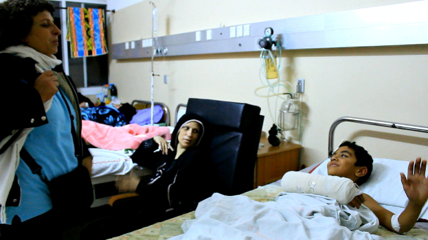 Ein Krankenhauszimmer mit mehreren Betten, von denen jedes von einem Patienten belegt ist, Infusionsflaschen auf Ständern zwischen den Betten und eine Frau mit einer Tasche in der Nähe eines der Patienten.