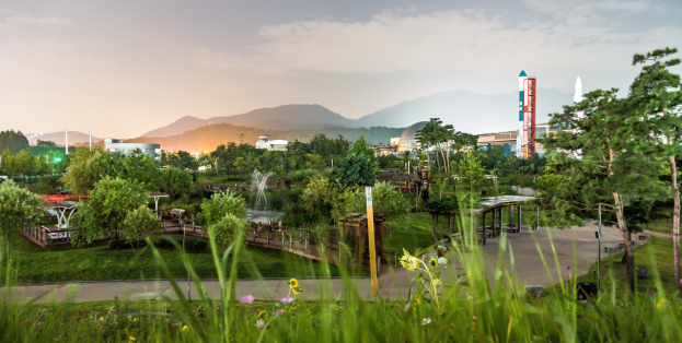 Ein Park mit grünem Gras, bunten Blumen, hohen Bäumen, einer Brücke, einem Springbrunnen, Laternenmasten und fernen Gebäuden, vor Bergen und einem klaren blauen Himmel.
