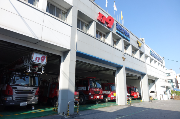 Feuerwache mit mehreren Feuerwehrautos davor, mit Fenstern, Säulen, Namensschildern und Fahnenmasten im Vordergrund, vor anderen Gebäuden und einem klaren blauen Himmel.
