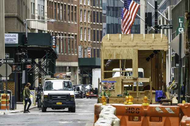 Eine Stadtstraße mit einem geparkten Lkw, fahrenden Fahrzeugen, Fußgängern auf dem Gehweg, Gebäuden im Hintergrund, einer Fahne auf der rechten Seite und Verkehrszeichen und Schilder an Pfosten entlang der Straße.