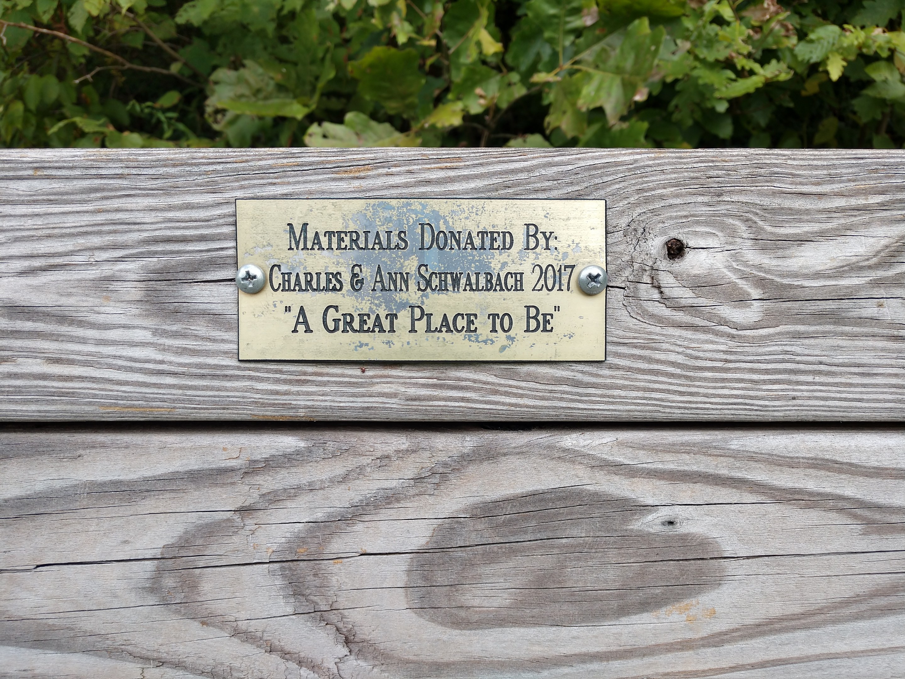 Holzbank mit einer Plakette, auf der "Materialien gespendet von Charles & Ann Schwabach 2017: Ein schöner Ort" steht, vor üppiger grüner Vegetation.