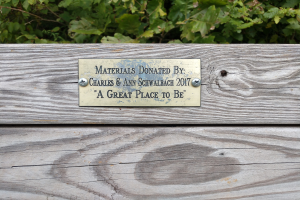 Holzbank mit einer Plakette, auf der "Materialien gespendet von Charles & Ann Schwabach 2017: Ein schöner Ort" steht, vor üppiger grüner Vegetation.