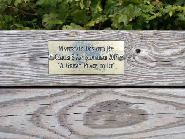 Holzbank mit einer Plakette, auf der "Materialien gespendet von Charles & Ann Schwabach 2017: Ein schöner Ort" steht, vor üppiger grüner Vegetation.