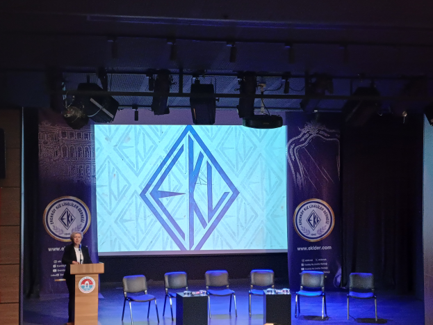 Eine Frau steht an einem Podium mit einem Mikrofon, spricht auf einer Konferenz mit Stühlen, Tischen und Banner auf der Bühne und wird von Lampen an der Decke beleuchtet.
