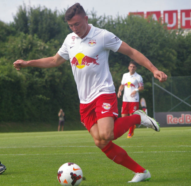 Ein professioneller Fußballspieler schießt einen Ball auf einem grünen Feld mit Bäumen und einem klaren blauen Himmel im Hintergrund, in der Nähe eines Schildes mit der Aufschrift "RB Leipzig."