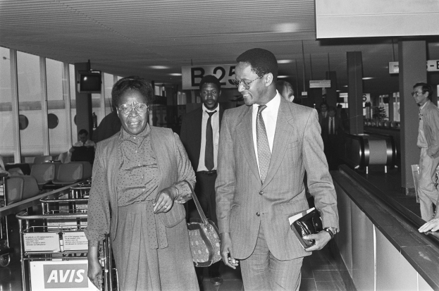 Schwarzes und weißes Foto eines Mannes in einem Anzug und einer Frau mit einer Handtasche, die durch einen Flughafen gehen, umgeben von anderen Reisenden.
