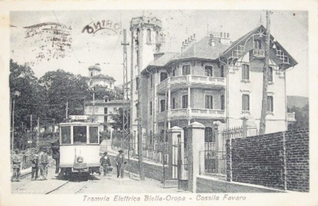 Ein altes Schwarz-Weiß-Foto einer Straßenbahn auf Schienen, umgeben von Gebäuden, Bäumen, Pfählen und Menschen, mit Text am unteren Rand.