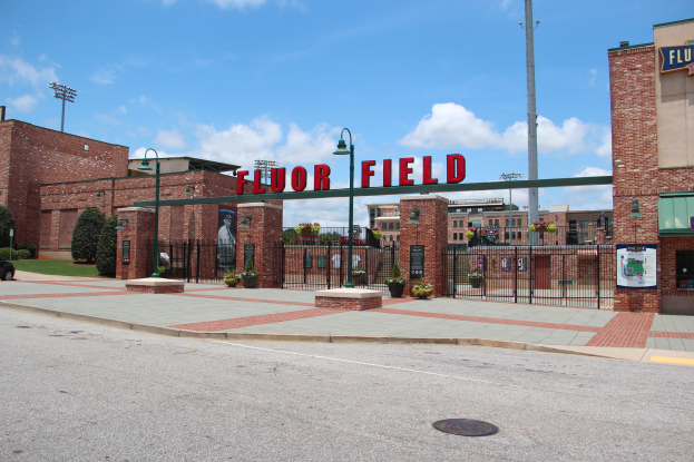 Der Eingang zu einem Baseballstadion mit einem Schild, das "Floor Field" lautet, umgeben von Gebäuden, Straßenlaternen, Grills, Namensschildern, Kraftfahrzeugen, Büschen, Bäumen und einem bewölkten Himmel im Hintergrund.