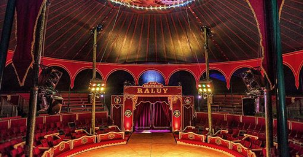 Das Innere eines großen Zirkuszeltes mit roten Sitzreihen, Stangen, Lichtern, einer Bühne, einem Vorhang, einem Namensschild und anderen Gegenständen, wahrscheinlich das historische Raluy Theatre.