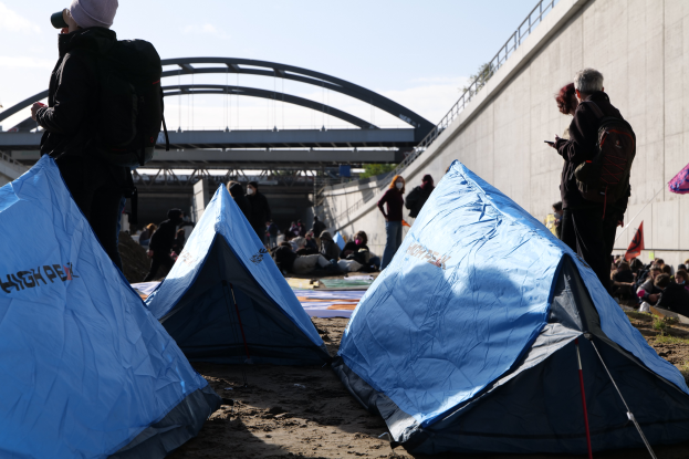Eine Gruppe von Menschen sitzt auf einem sandigen Strand in der Nähe von Zelten, mit einer Wand auf der rechten Seite und einer Brücke im Hintergrund, haltend Gegenstände und tragend Taschen während einer Klimawandel-Demonstration unter einem bewölkten Himmel.