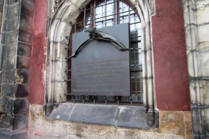 Steinbau mit Fenster und Holocaust-Gedenktafel an der Seite, mit eingravierter Inschrift.