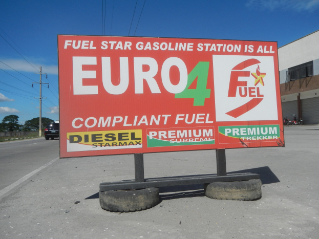 Eine Tankstelle mit einem "Euro4 Fuel"-Schild im Vordergrund, Fahrzeuge auf der Straße, Strommasten mit Drähten, Bäume und ein Gebäude im Hintergrund unter einem sichtbaren Himmel.