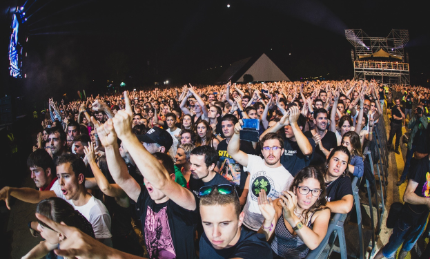 Eine große Menschenmenge auf einem Musikfestival mit erhobenen Händen, einige sitzen auf Stühlen und andere stehen, begrenzt von einem Metallzaun auf der rechten Seite, mit Gebäuden, Lichtern und einem dunklen Himmel im Hintergrund.