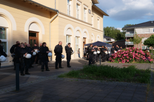 Gruppe von Polizisten in Uniform vor einem Gebäude mit Fenstern und Türen stehend, Helme haltend, neben einem Zelt, umgeben von Grün, einem Straßenmast, Bäumen und einem bewölkten Himmel.