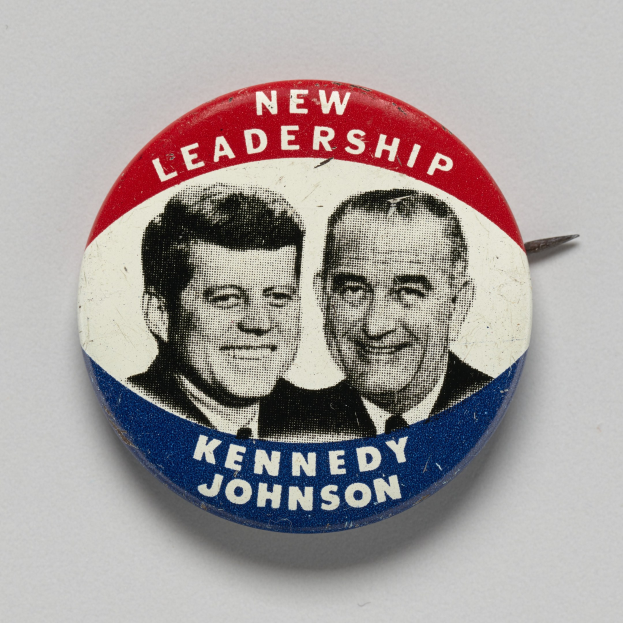 Weißer Ansteckknopf mit zwei Fotos von Kennedy und Johnson, beschriftet mit "Neue Führung", auf einer glatten Oberfläche.