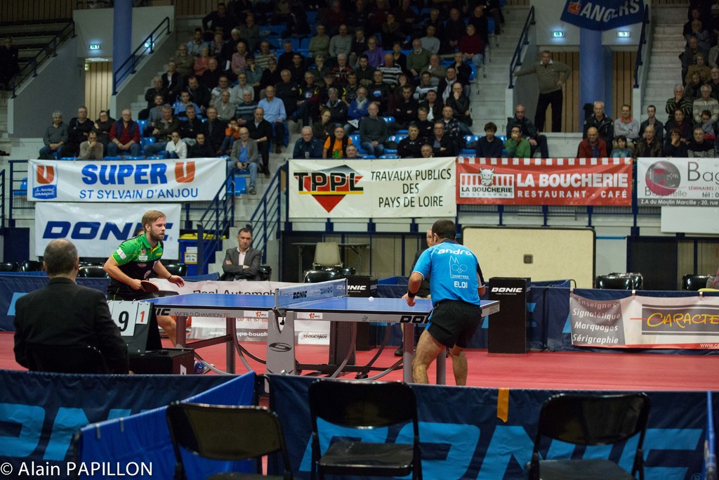 Zwei Männer spielen drinnen Tischtennis, mit mehreren Zuschauern, die stehen und sitzen, leeren Stühlen und Werbeplakaten, mit Text in der linken unteren Ecke.