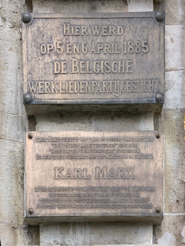 Eine Metalltafel an einer Gebäudewand mit der Aufschrift "Karl Marx" in fetter schwarzer Schrift.
