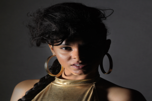 Eine Frau in einem goldenen Kleid und großen Creolen steht selbstbewusst vor einer Wand, ihre glatte Bob-Frisur ist ordentlich gestylt, trägt dezentes Make-up mit pinkem Rouge und glänzenden Lippen, schaut direkt in die Kamera mit einem entschlossenen Gesichtsausdruck.