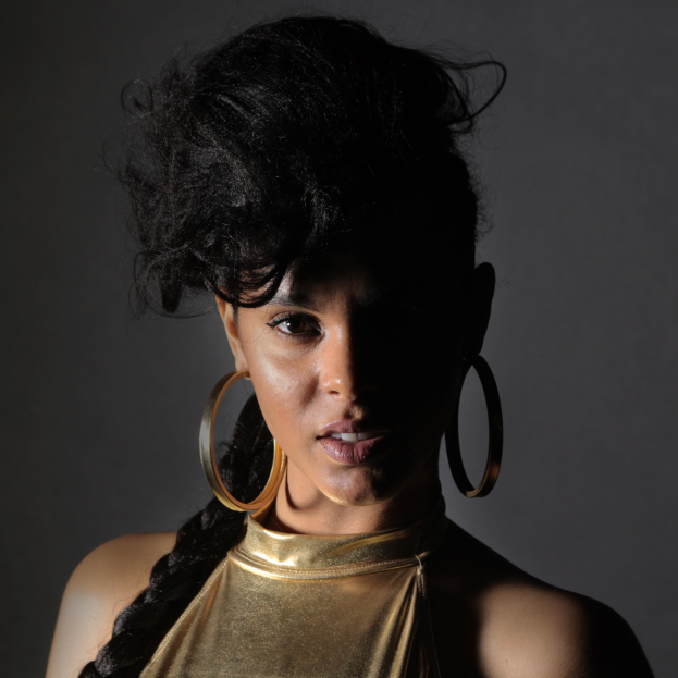 Eine Frau in einem goldenen Kleid und großen Creolen steht selbstbewusst vor einer Wand, ihre glatte Bob-Frisur ist ordentlich gestylt, trägt dezentes Make-up mit pinkem Rouge und glänzenden Lippen, schaut direkt in die Kamera mit einem entschlossenen Gesichtsausdruck.